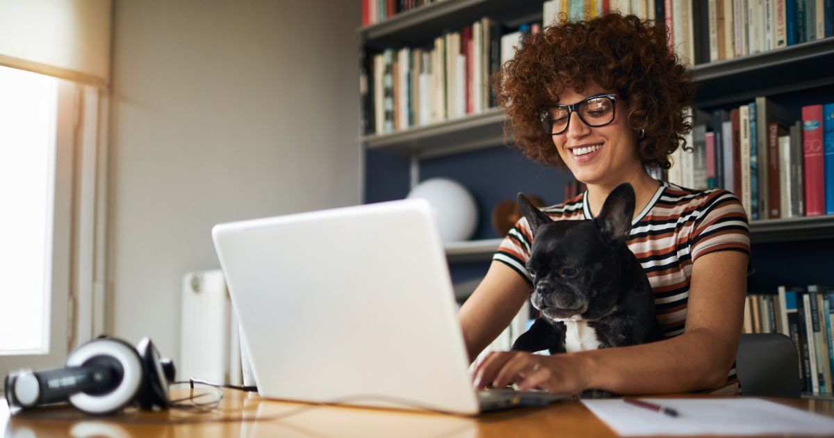 Woman working from home on a laptop with a dog, surrounded by bookshelves, illustrating the benefits of remote work and a comfortable workspace.