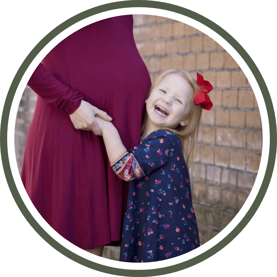 img-happy-moms-1@2x Pregnant woman in maroon dress with young girl in floral dress, both smiling and embracing, symbolizing prenatal and pediatric care at Foundation Family Chiropractic.