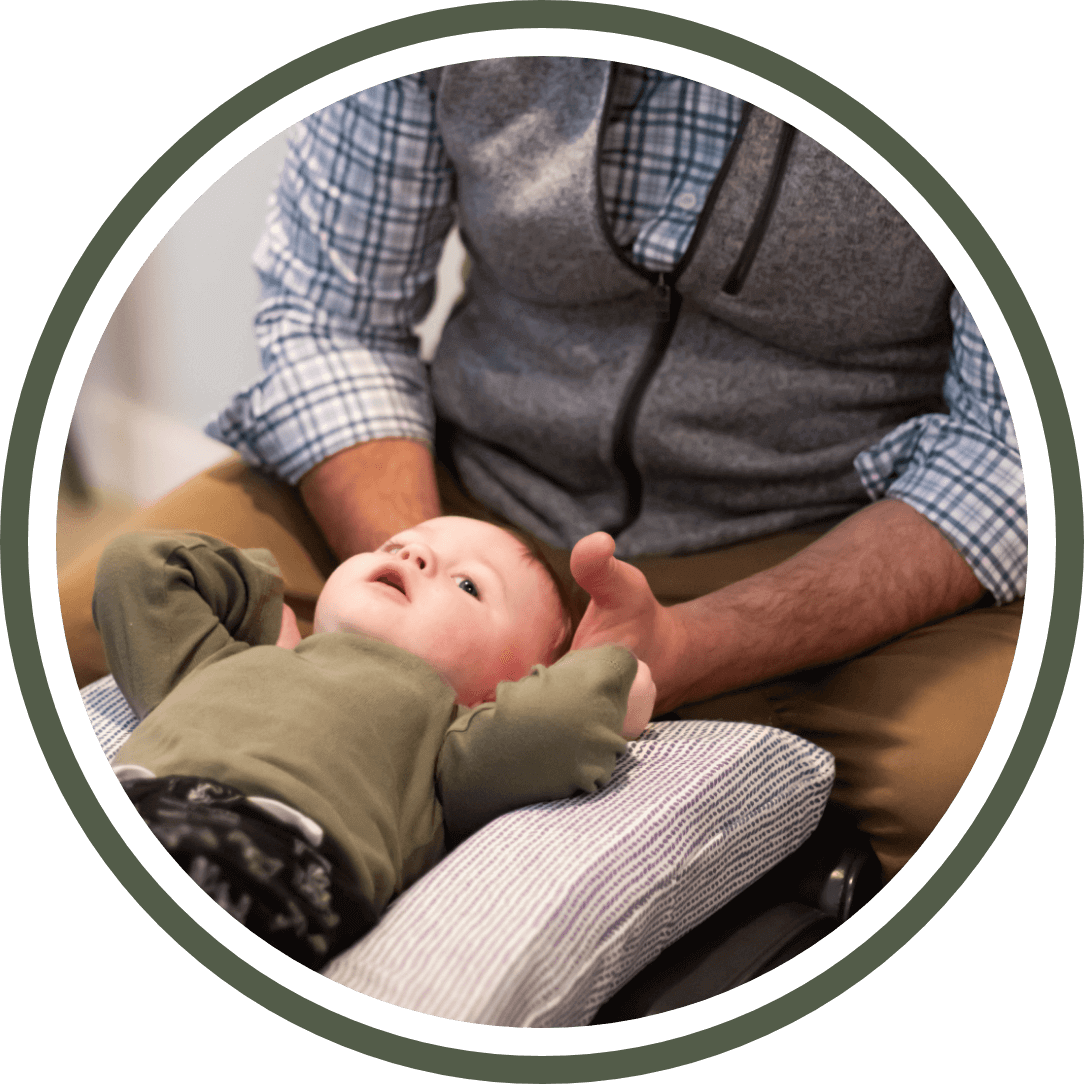 img-happy-moms-2@2x Baby receiving chiropractic care from practitioner in a warm, family-friendly setting, emphasizing postnatal health and wellness.