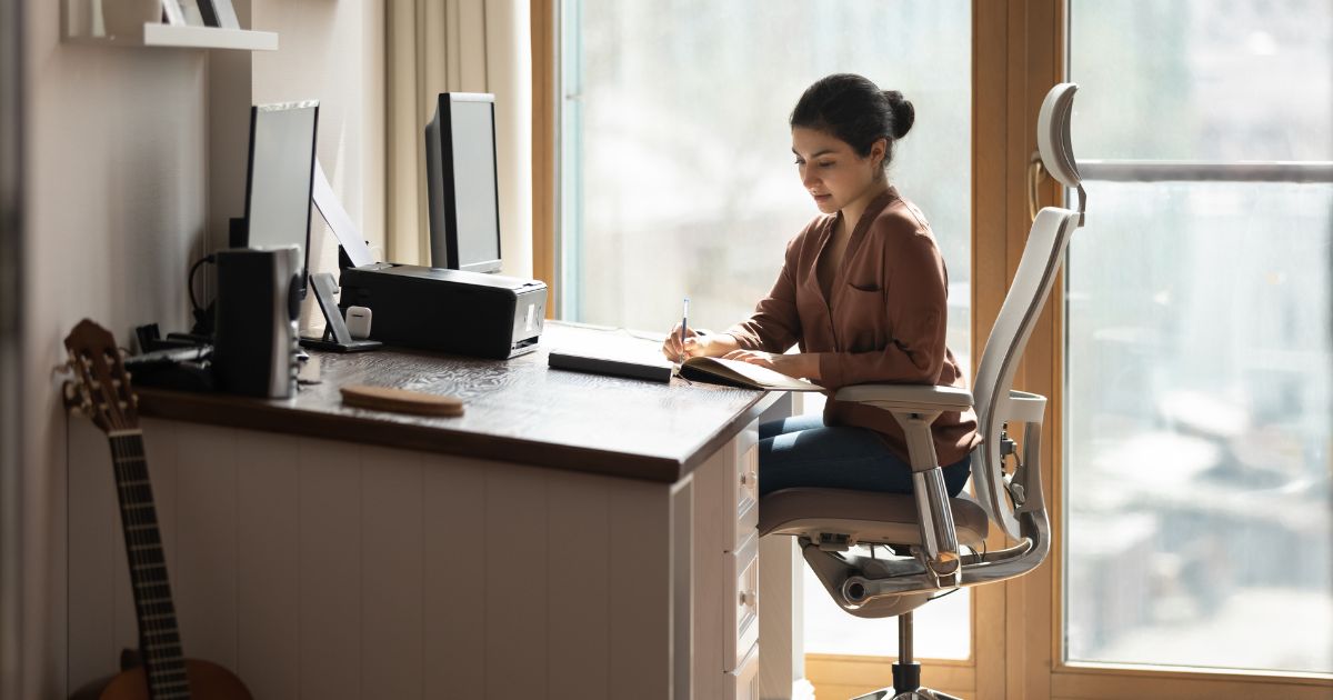 Woman sitting at a desk in an ergonomic chair, writing in a notebook, with dual computer monitors and a printer, emphasizing workplace ergonomics and health.