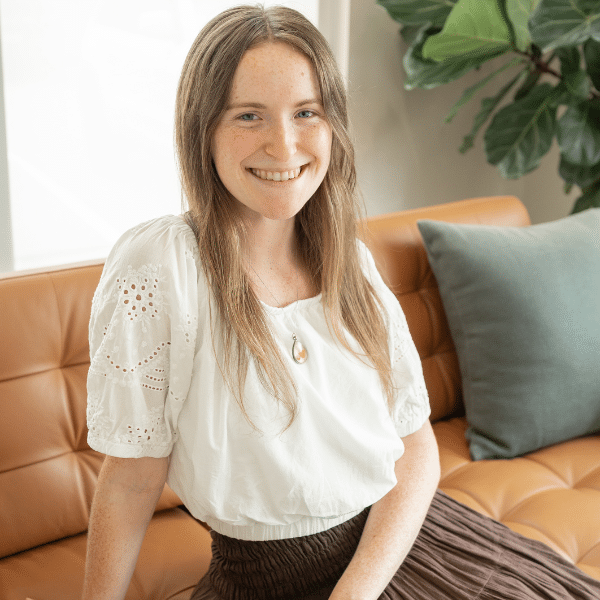 Smiling young woman with long hair wearing a white blouse, seated on a brown couch with green cushions, representing a chiropractic assistant at Foundation Family Chiropractic.
