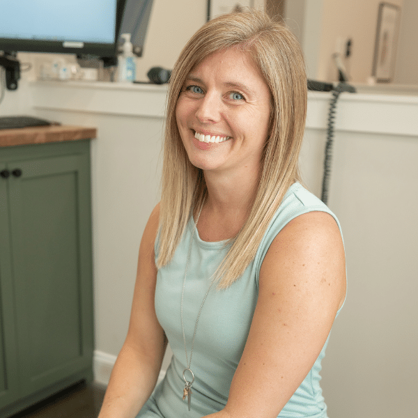 Chiropractic Assistant Amanda smiling in a light blue shirt, seated in a wellness clinic environment with a warm, inviting atmosphere.