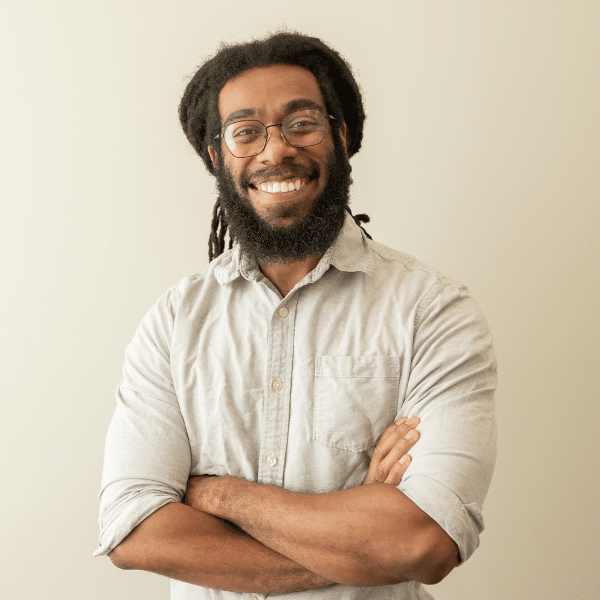Barrett, Chiropractic Assistant, smiling with arms crossed, wearing a light gray shirt and glasses, against a neutral background, reflecting a warm and welcoming demeanor.