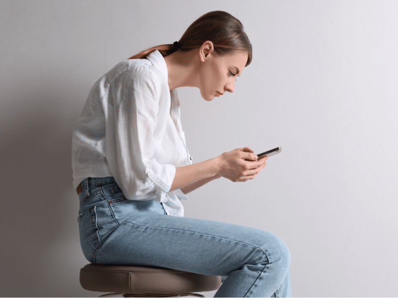 Woman sitting with poor posture while using a smartphone, illustrating the impact of daily activities on spinal health and posture.